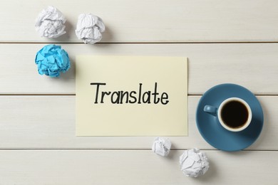 Photo of Card with word Translate, crumpled paper balls and cup of coffee on white wooden table, flat lay