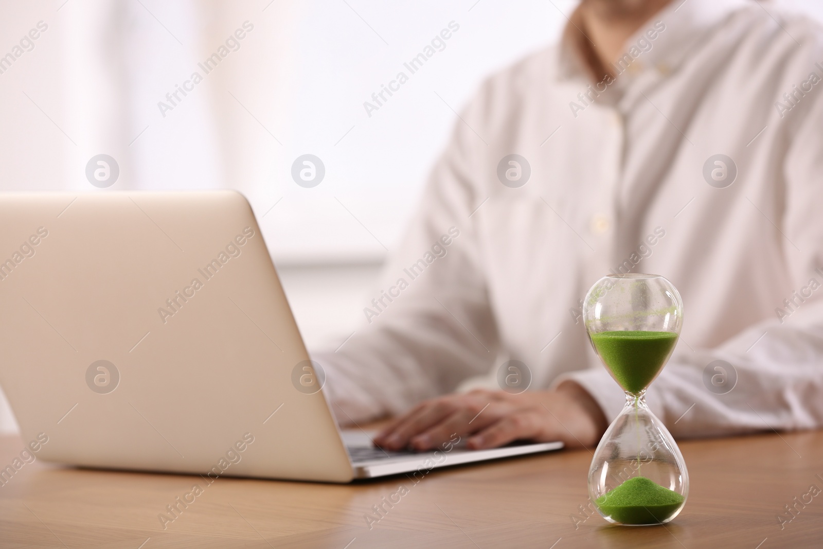 Photo of Hourglass with flowing sand on wooden table, selective focus. Man using laptop indoors