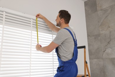 Photo of Worker in uniform using measuring tape while installing horizontal window blinds indoors
