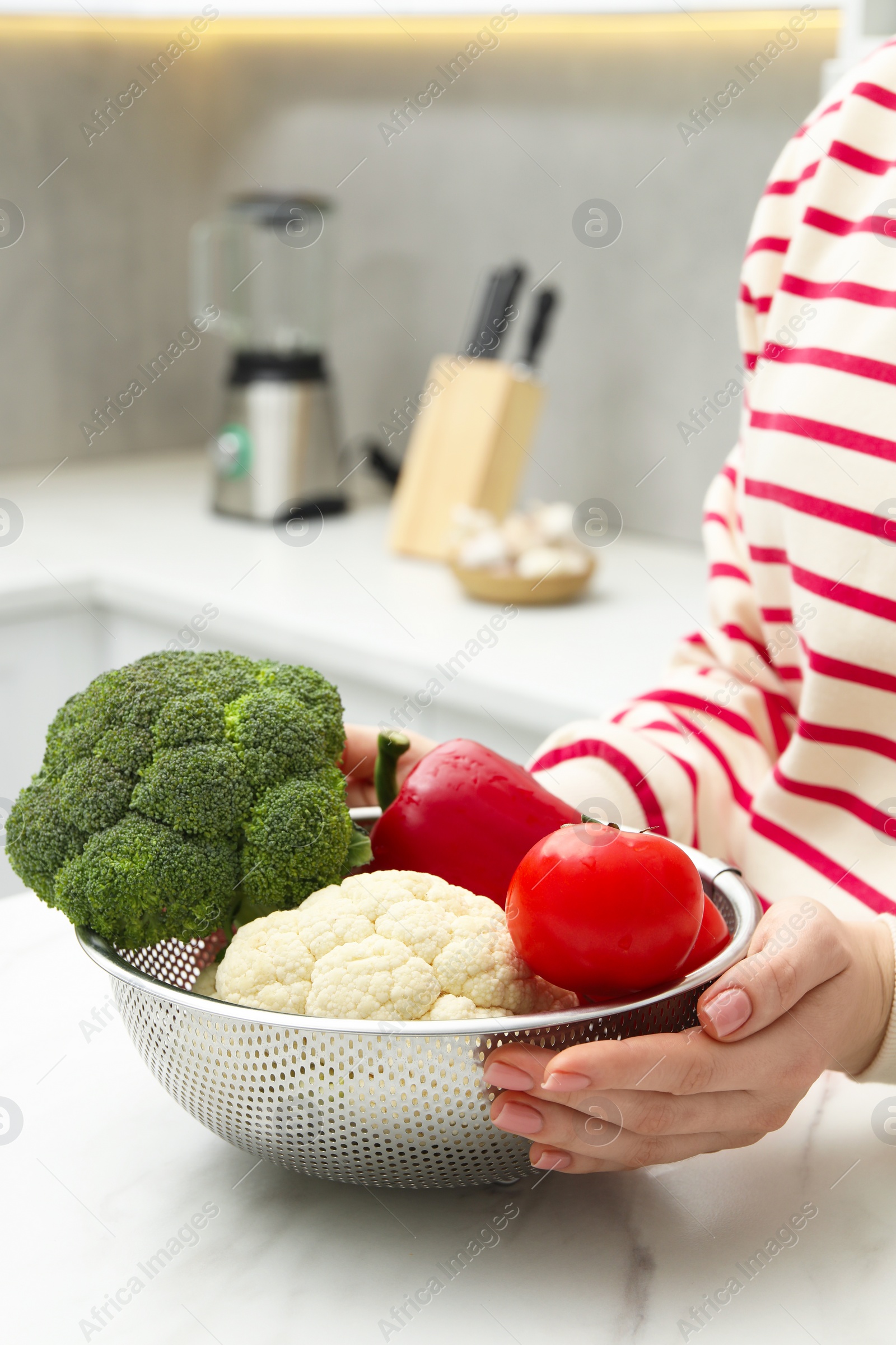 Photo of Woman holding colander with fresh vegetables at white marble table in kitchen, closeup