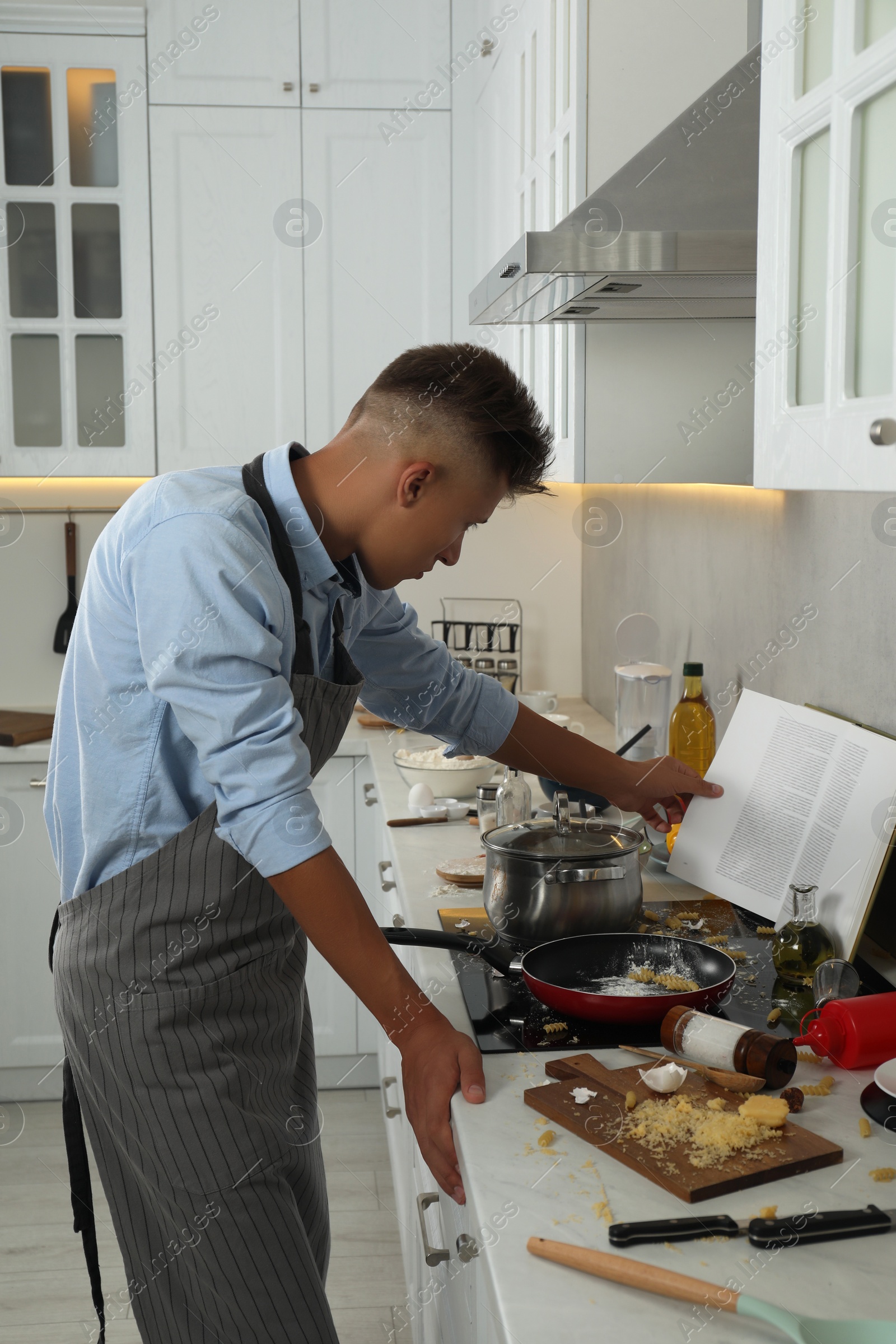 Photo of Man reading cookery book in messy kitchen. Many dirty dishware and utensils on stove and countertop