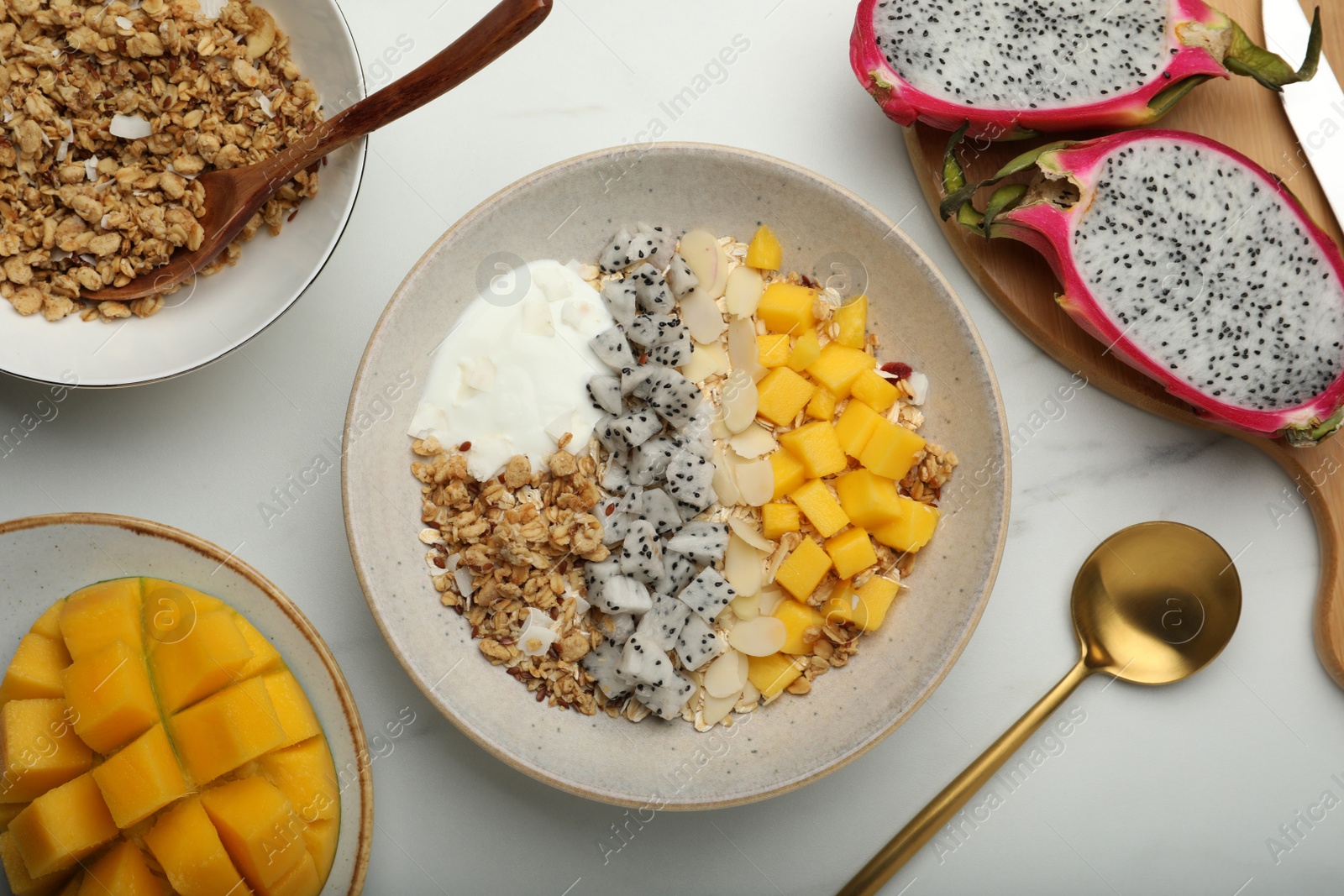 Photo of Bowl of granola with pitahaya, mango and yogurt near ingredients on white marble table, flat lay
