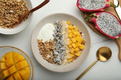 Photo of Bowl of granola with pitahaya, mango and yogurt near ingredients on white marble table, flat lay