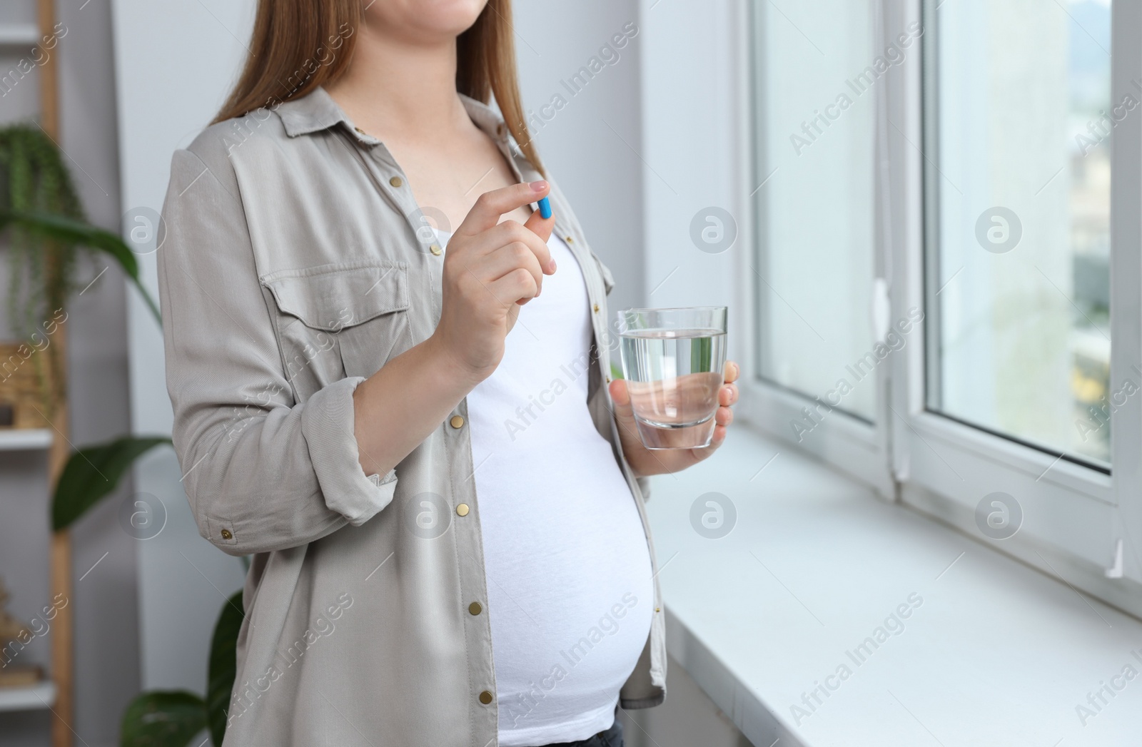 Photo of Pregnant woman holding pill and glass with water near window at home, closeup