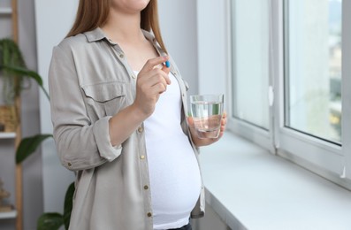 Pregnant woman holding pill and glass with water near window at home, closeup