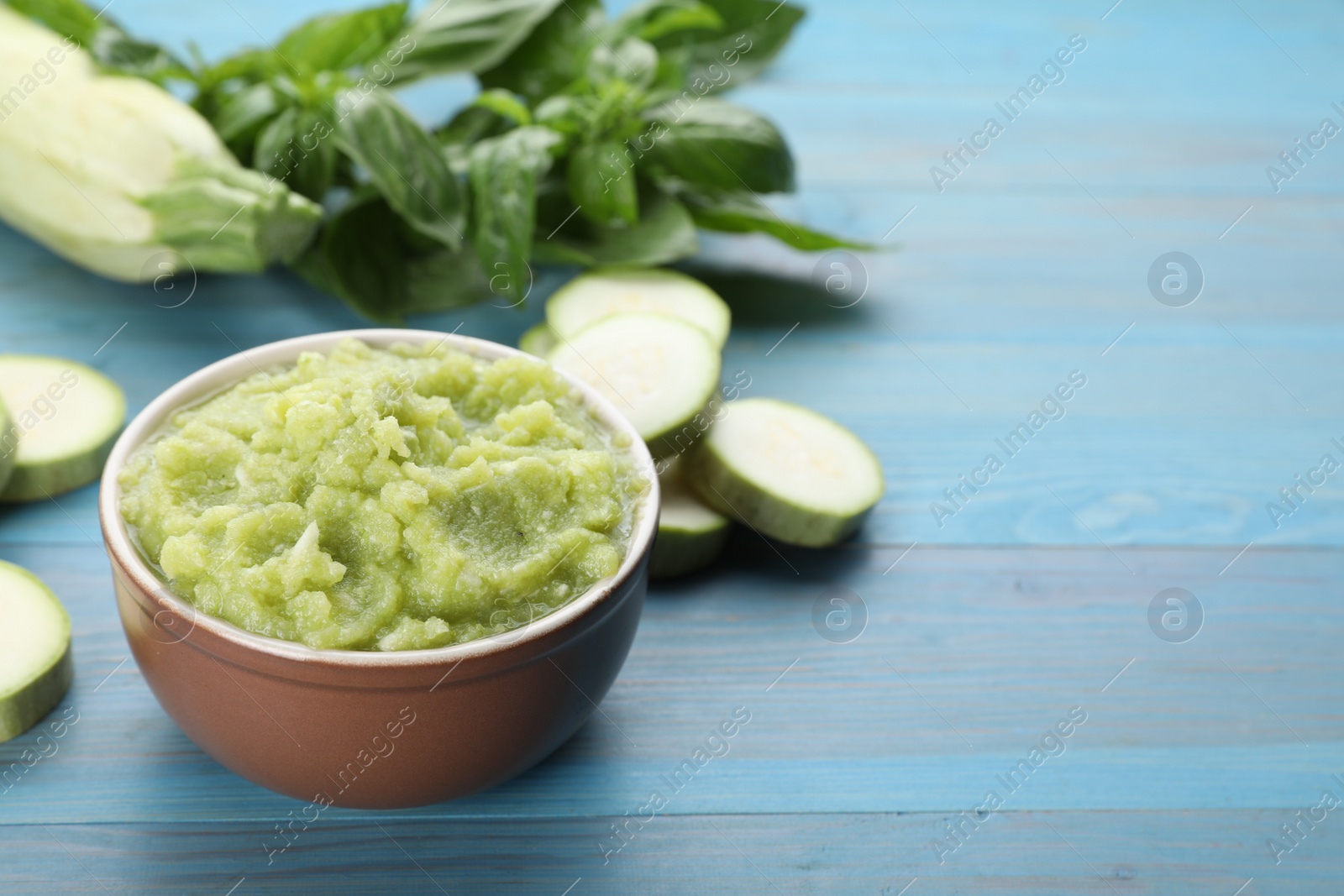 Photo of Bowl with green tasty puree, zucchini and basil on light blue wooden table. Space for text