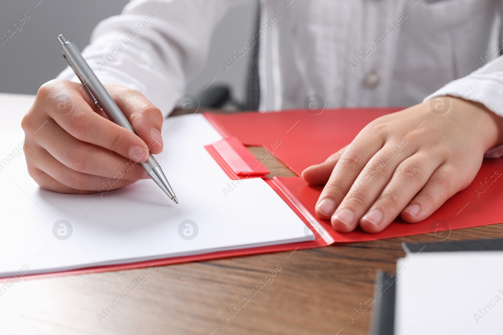 Photo of Woman writing on sheet of paper in red folder at wooden table in office, closeup