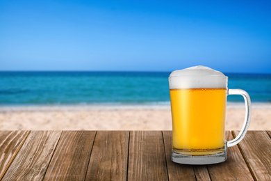 Image of Cold beer on wooden desk against blurred sea and sandy beach. Space for text