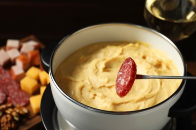 Photo of Dipping piece of sausage into fondue pot with melted cheese at table, closeup