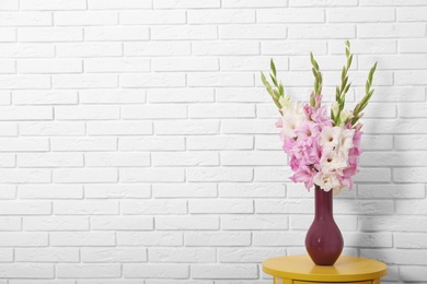 Vase with beautiful gladiolus flowers on wooden table against white brick wall. Space for text