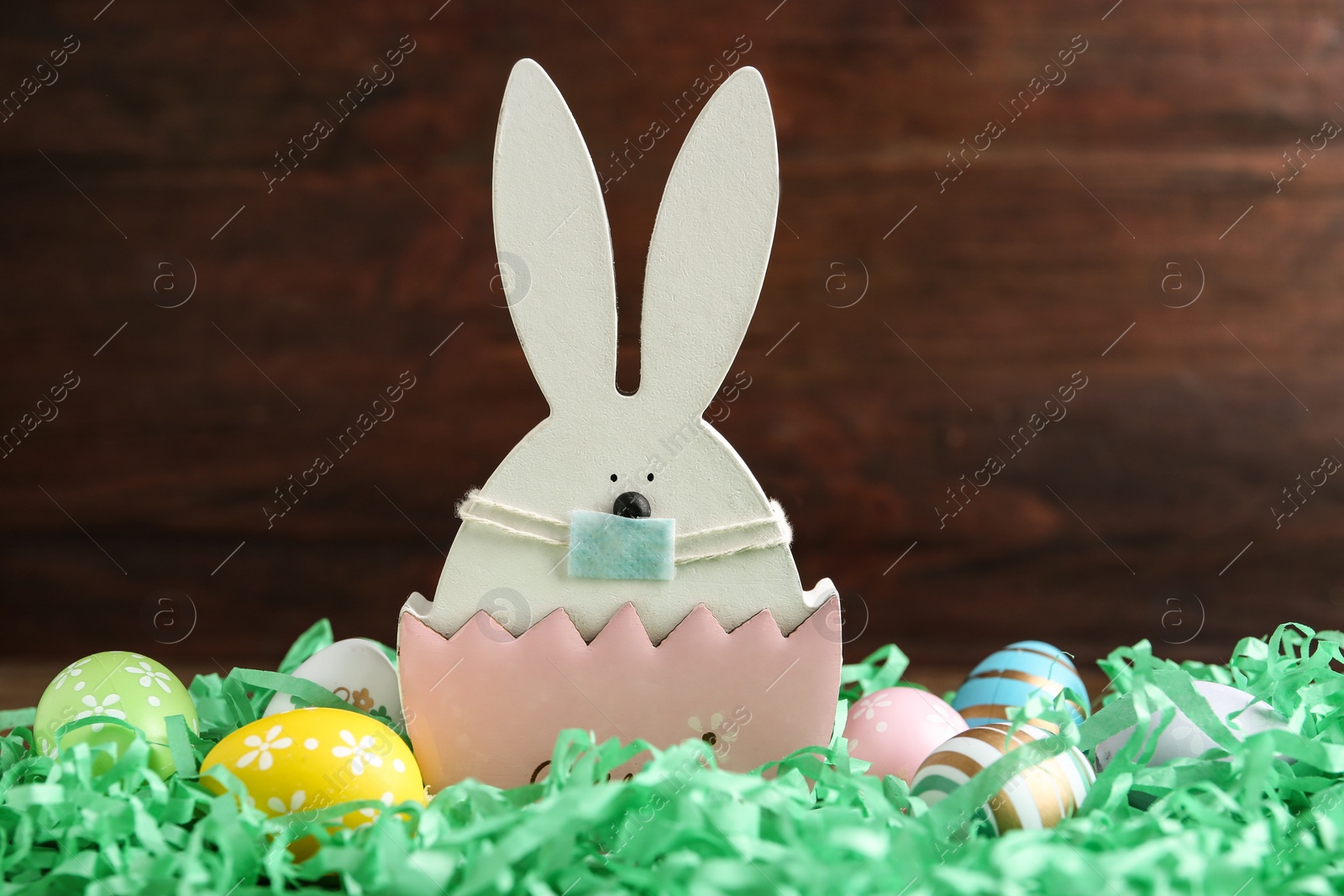 Photo of Cute bunny figure in protective mask and dyed eggs on wooden background. Easter holiday during COVID-19 quarantine