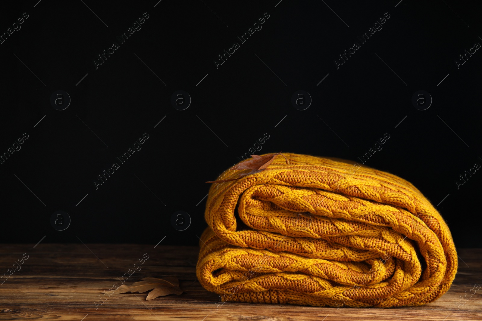 Photo of Orange knitted plaid with dry leaves on wooden table. Space for text