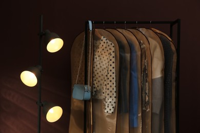 Photo of Garment bags with clothes hanging on rack near lamp and brown wall