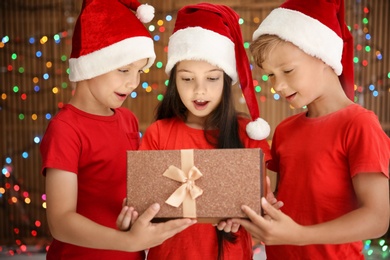 Photo of Cute little children in Santa hats opening Christmas gift box on blurred lights background