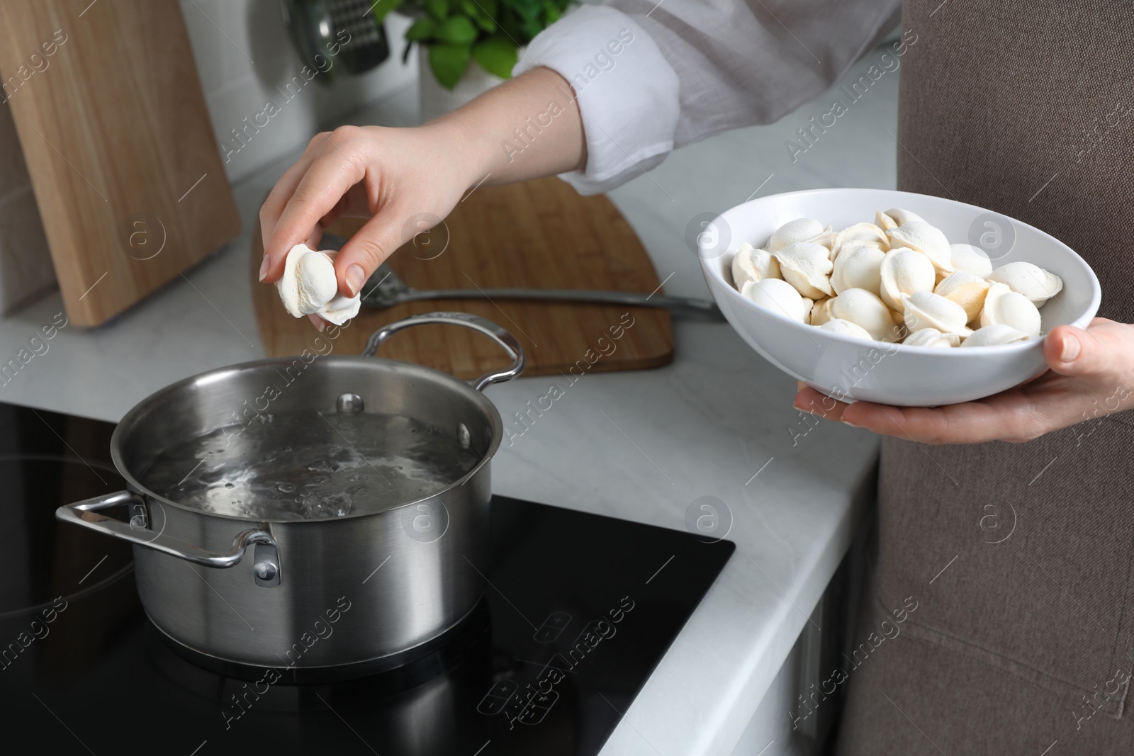 Photo of Woman putting frozen dumplings into saucepan with boiling water on cooktop in kitchen, closeup