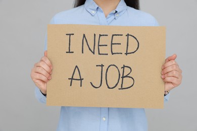 Photo of Young unemployed woman holding sign with phrase I Need A Job on grey background, closeup