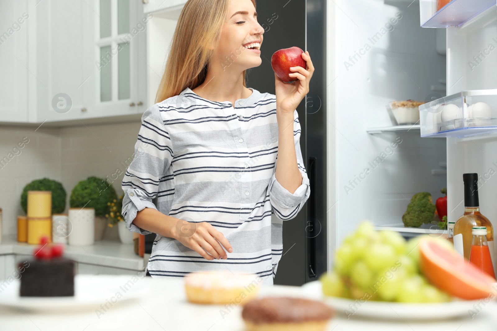 Photo of Concept of choice between healthy and junk food. Woman eating fresh apple near refrigerator in kitchen