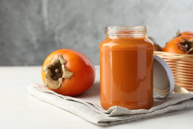 Photo of Delicious persimmon jam in glass jar and fresh fruits on white wooden table