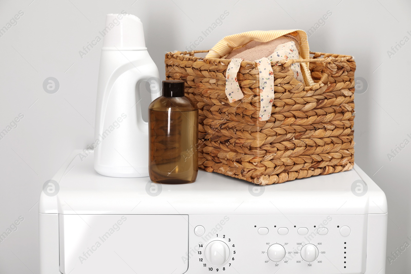 Photo of Baby clothes in wicker basket and laundry detergents on washing machine near light wall