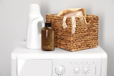 Photo of Baby clothes in wicker basket and laundry detergents on washing machine near light wall