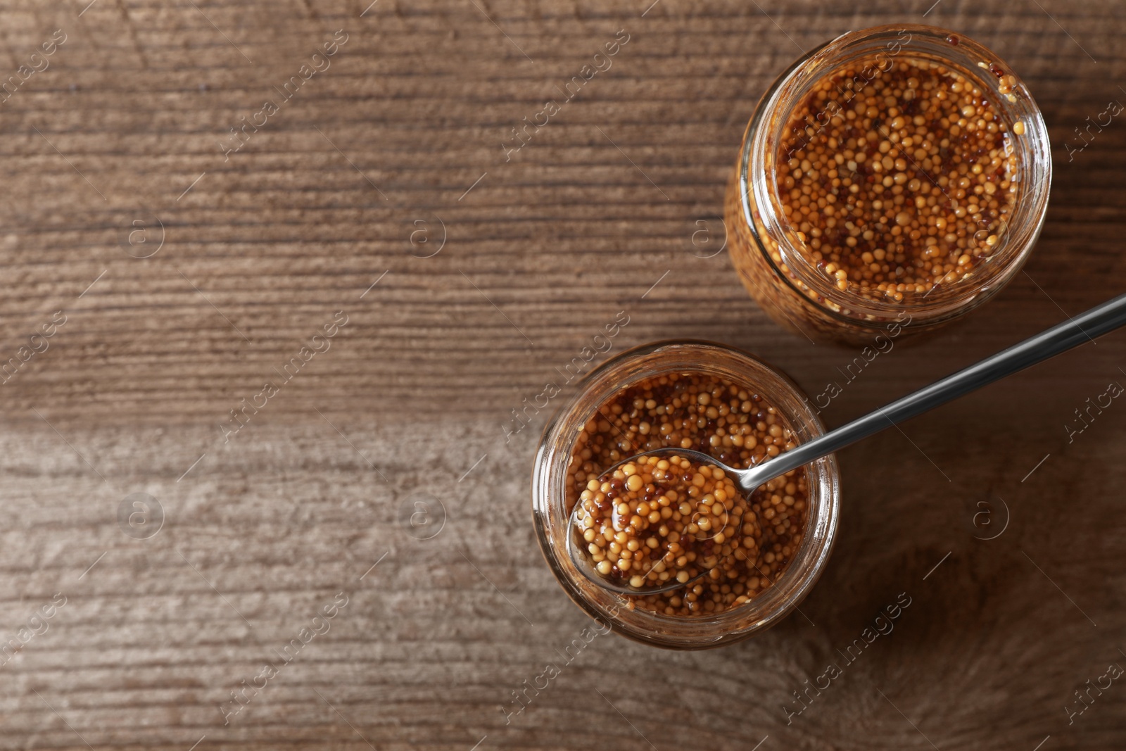 Photo of Jars and spoon of whole grain mustard on wooden table, flat lay. Space for text