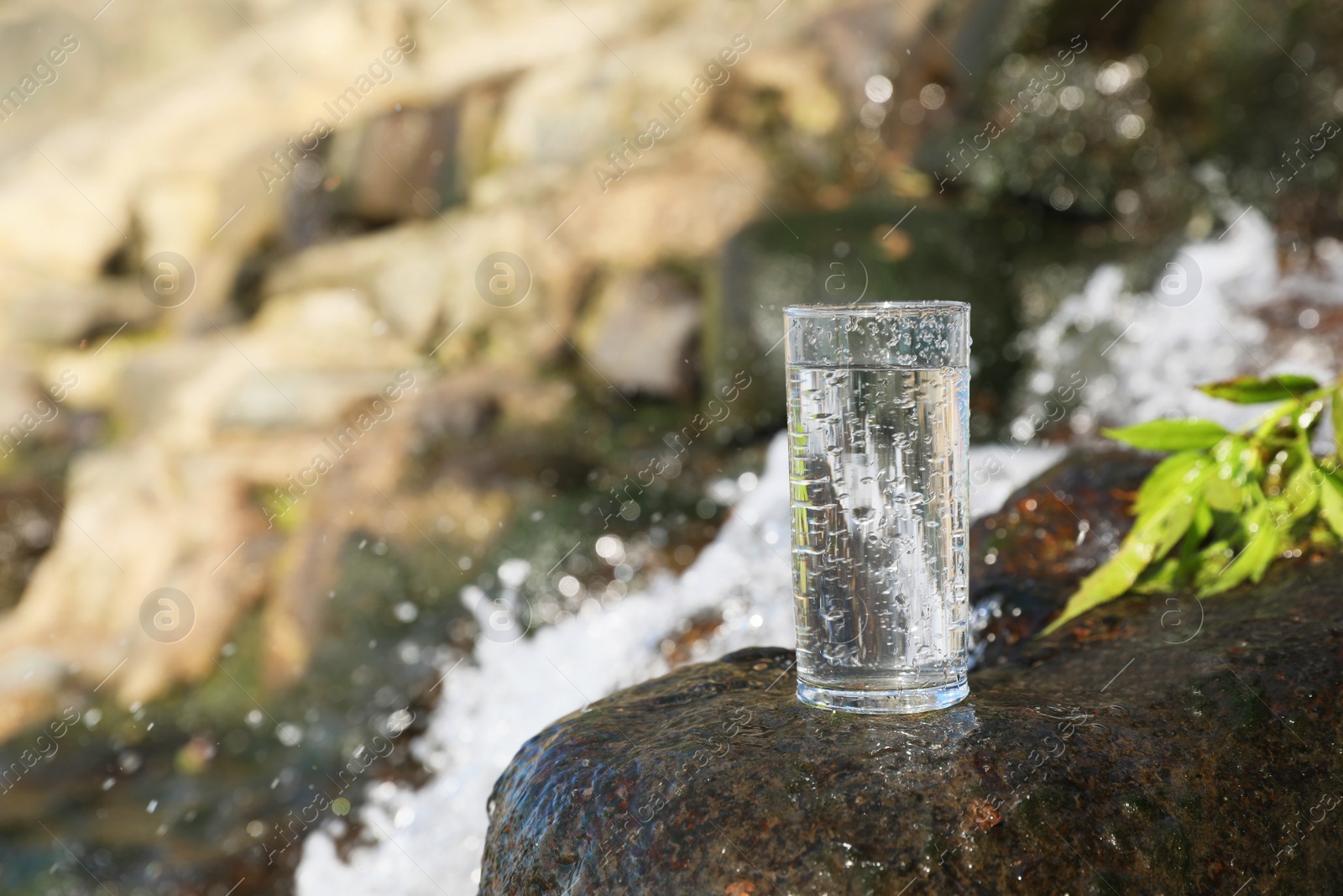 Photo of Wet glass of water on rocks near flowing stream outdoors, space for text