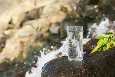 Photo of Wet glass of water on rocks near flowing stream outdoors, space for text