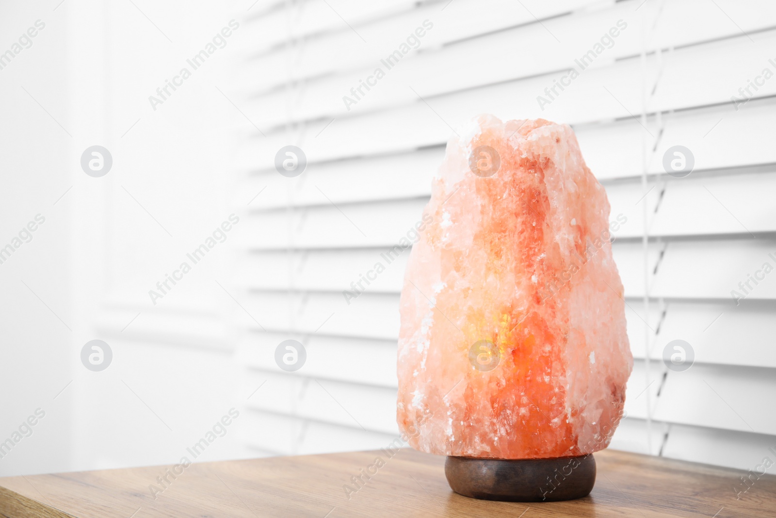 Photo of Himalayan salt lamp on wooden table near window blinds. Space for text