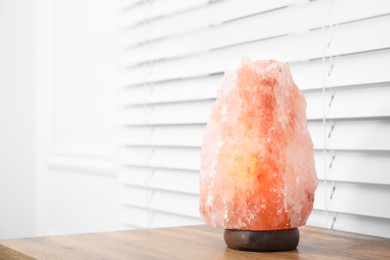 Photo of Himalayan salt lamp on wooden table near window blinds. Space for text