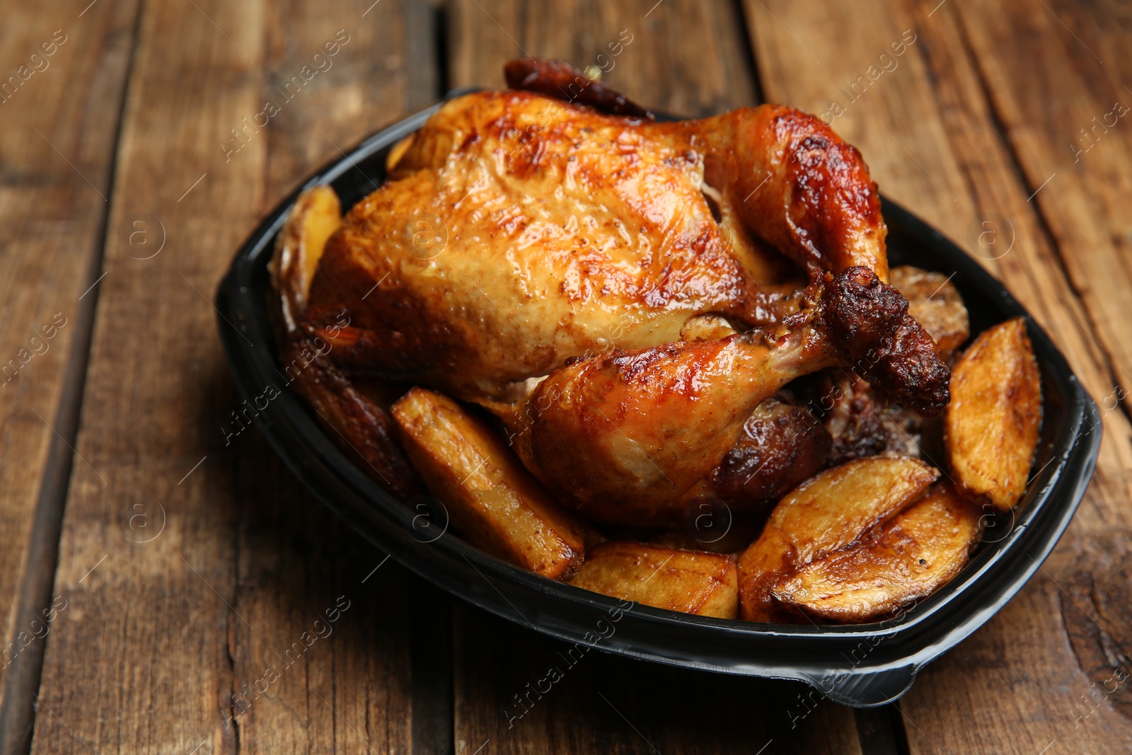 Photo of Delicious grilled whole chicken with potato in plastic container on wooden table. Food delivery service