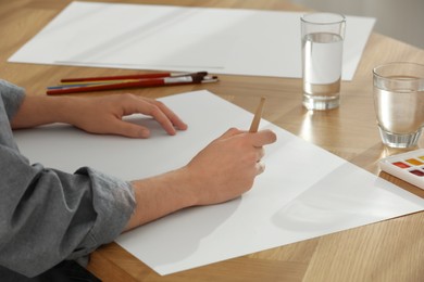 Photo of Man painting with watercolor on blank paper at wooden table, closeup