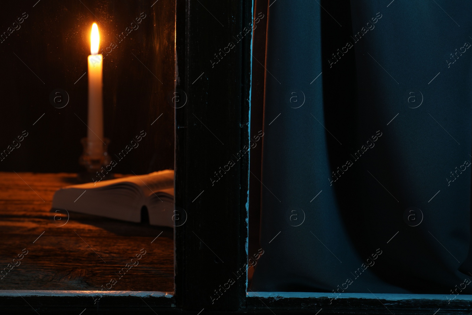 Photo of Burning candle and Bible on wooden table at night, view through window