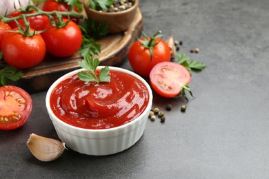 Photo of Delicious ketchup in bowl, tomatoes, parsley and garlic on grey table, closeup. Space for text