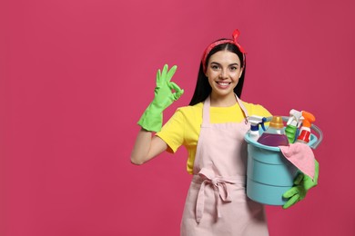Photo of Young housewife holding bucket with cleaning supplies on pink background, space for text