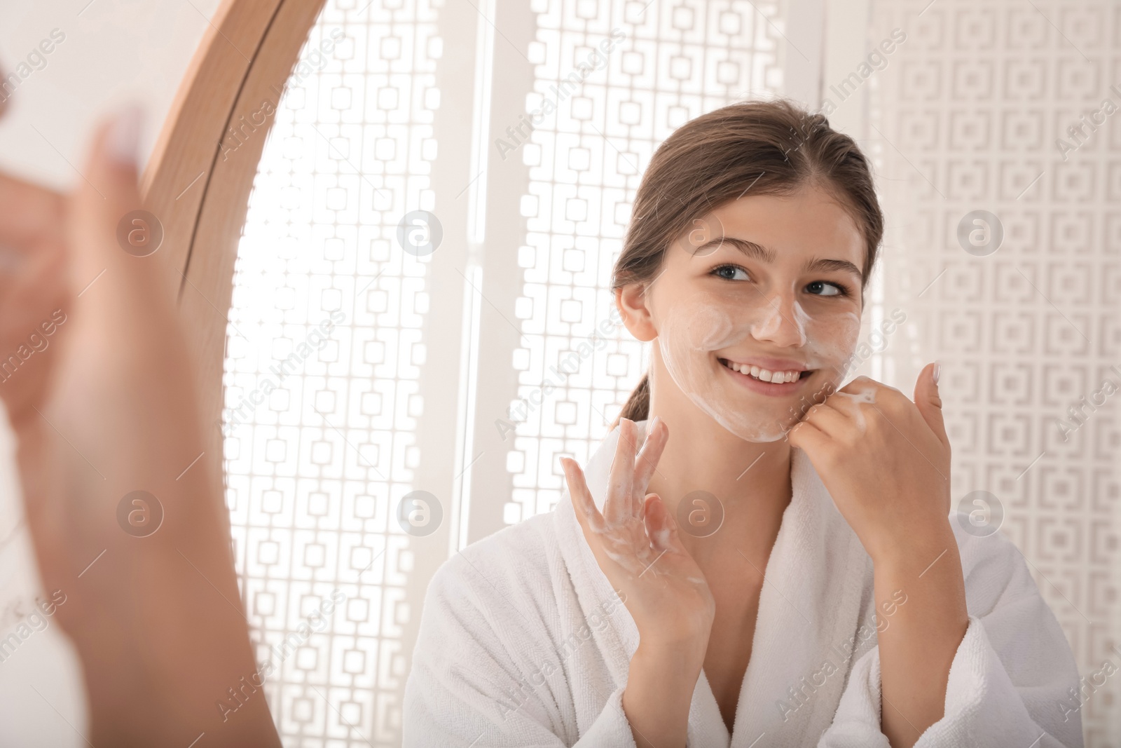 Photo of Beautiful teenage girl applying cleansing foam onto face near mirror at home. Skin care cosmetic