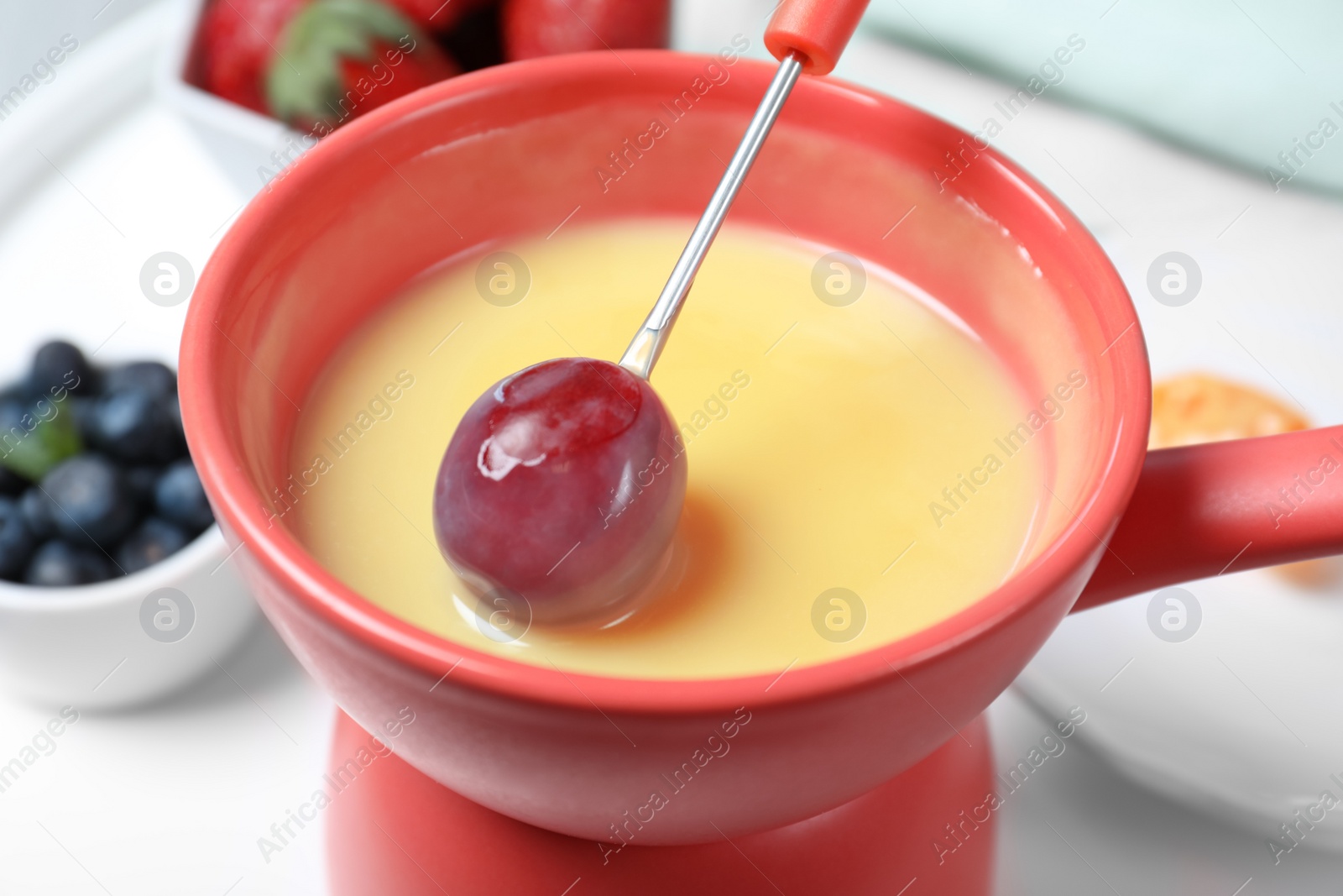 Photo of Dipping grape into pot with white chocolate fondue on table, closeup