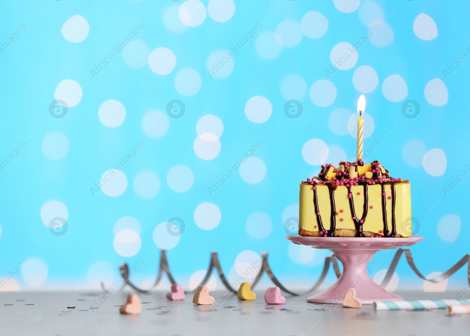 Photo of Piece of delicious birthday cake with burning candle on table against festive lights. Space for text