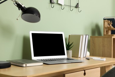Modern laptop, books, lamp and stationery on wooden desk near green wall. Home office
