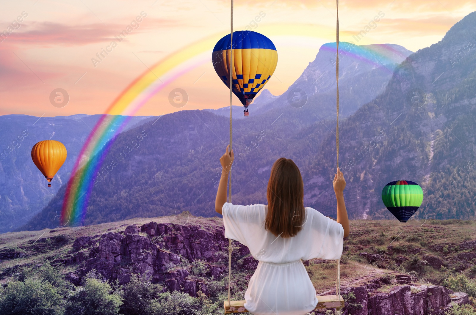 Image of Dream world. Young woman swinging over mountains, hot air balloons in sunset sky on background