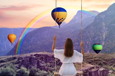 Dream world. Young woman swinging over mountains, hot air balloons in sunset sky on background