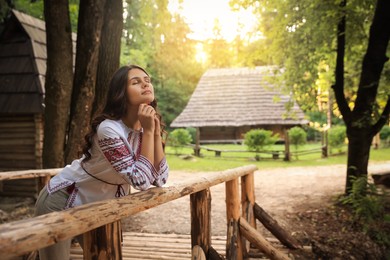 Photo of Beautiful woman wearing embroidered shirt near wooden railing in countryside, space for text. Ukrainian national clothes