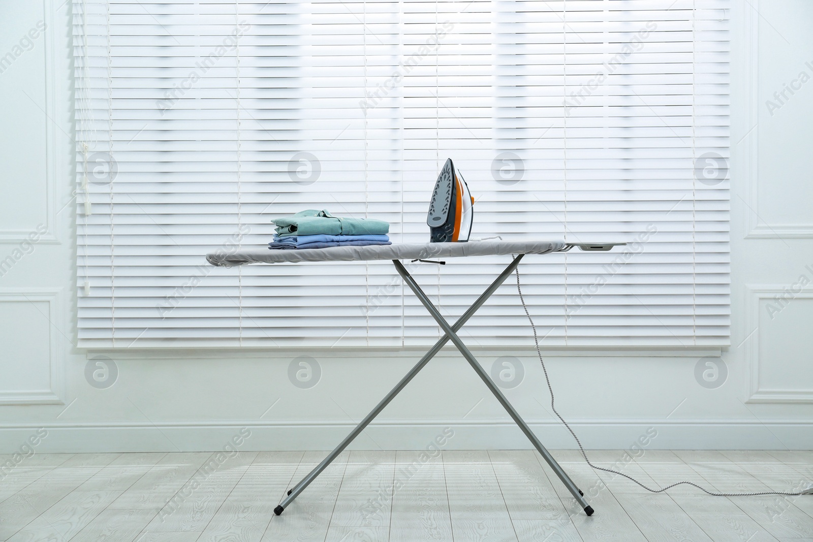 Photo of Modern iron and clean shirts on board near white blinds indoors