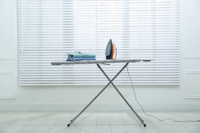 Modern iron and clean shirts on board near white blinds indoors