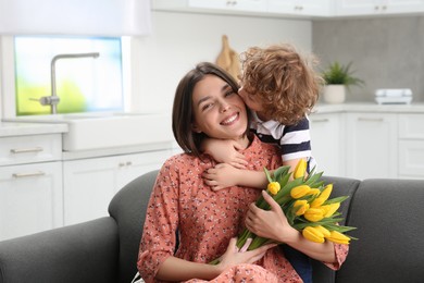 Photo of Little son kissing and congratulating his mom with Mother`s day at home. Woman holding bouquet of yellow tulips
