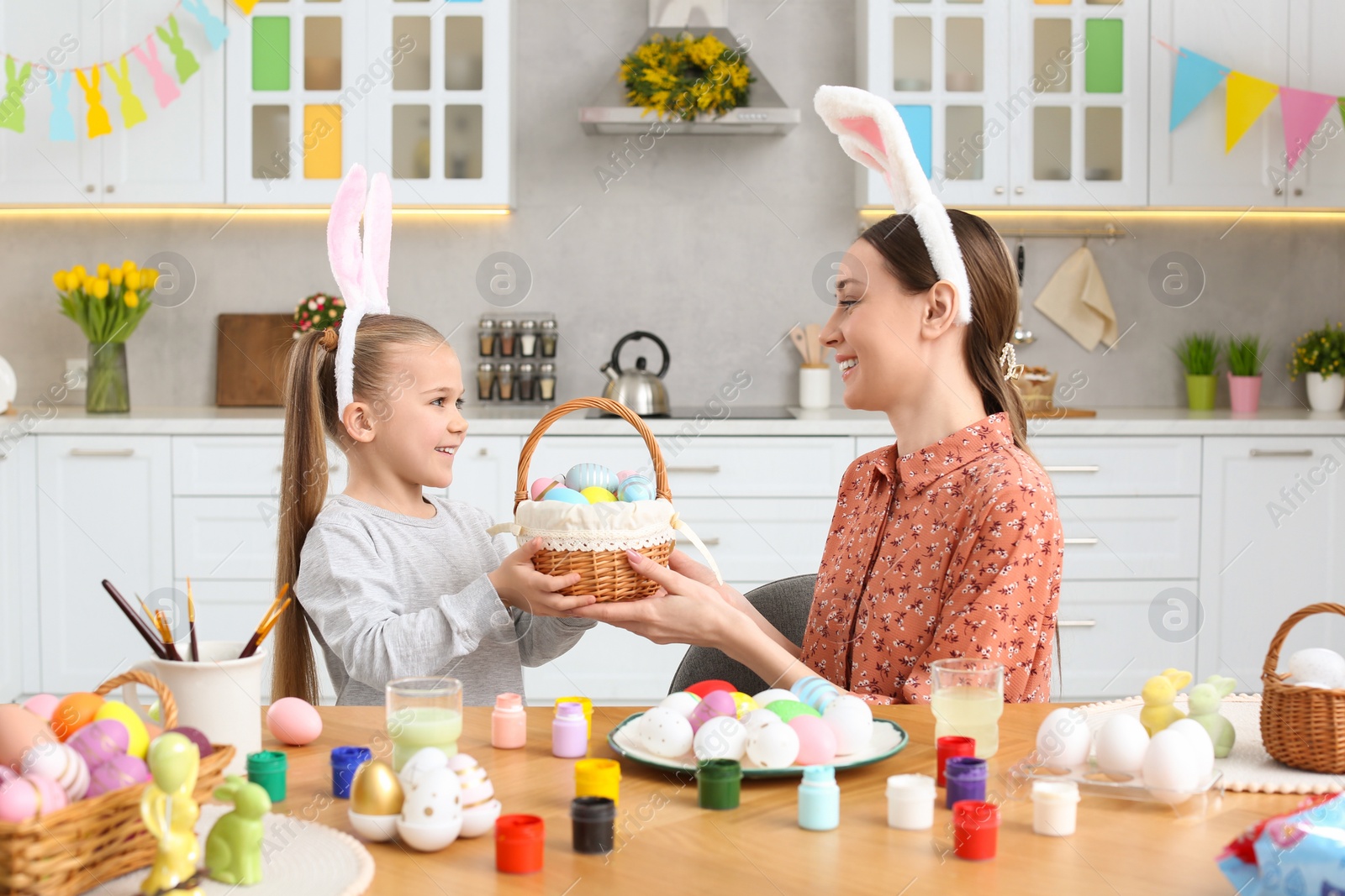 Photo of Mother and her cute daughter with wicker basket full of Easter eggs in kitchen