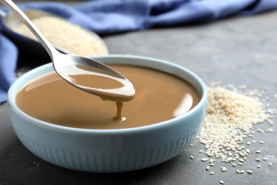 Photo of Spoon of tasty sesame paste above bowl on grey table, closeup