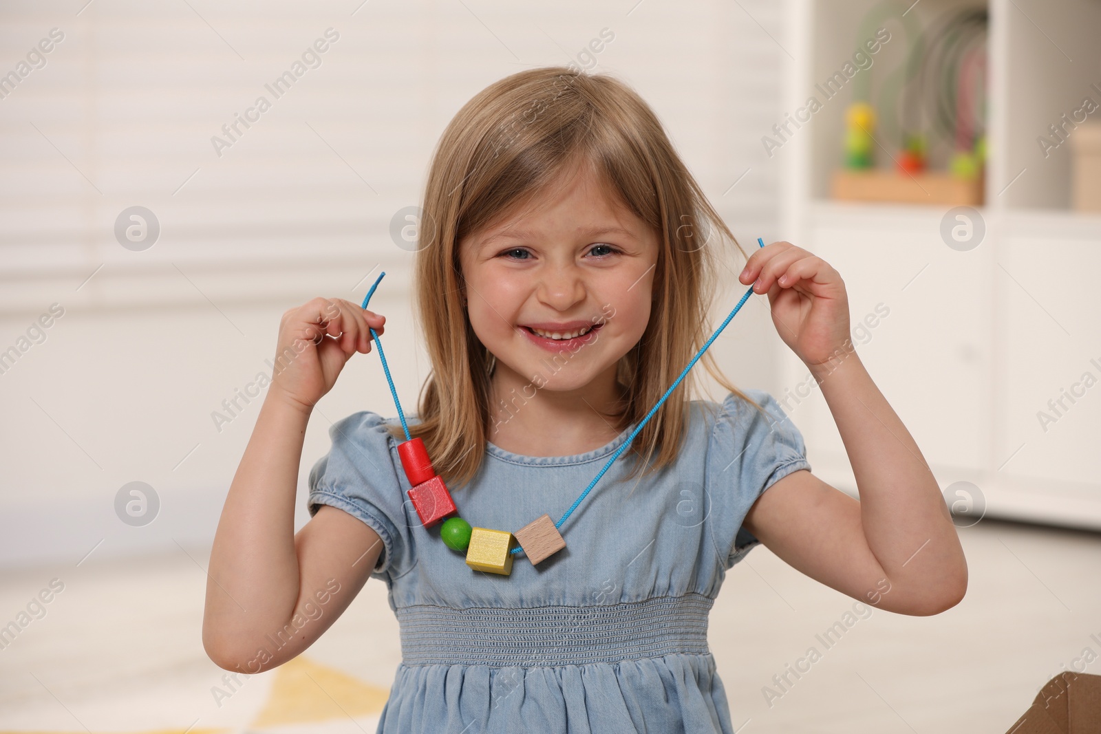 Photo of Cute little girl playing with wooden pieces and string for threading activity indoors. Child's toy