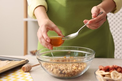 Photo of Making granola. Woman adding honey into bowl with mixture of oat flakes and other ingredients at light marble table in kitchen, closeup