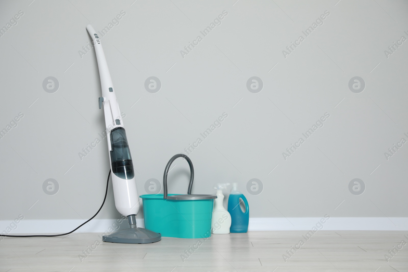 Photo of Modern steam mop and bucket with different cleaning supplies on floor near grey wall, space for text