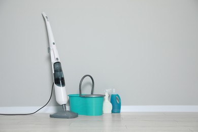 Modern steam mop and bucket with different cleaning supplies on floor near grey wall, space for text